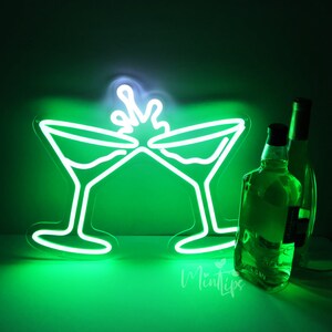 May include: A bright green neon sign depicting two martini glasses clinking together with a splash effect above them. Next to the glowing sign are two clear glass bottles, one positioned slightly behind the other, reflecting the green light.