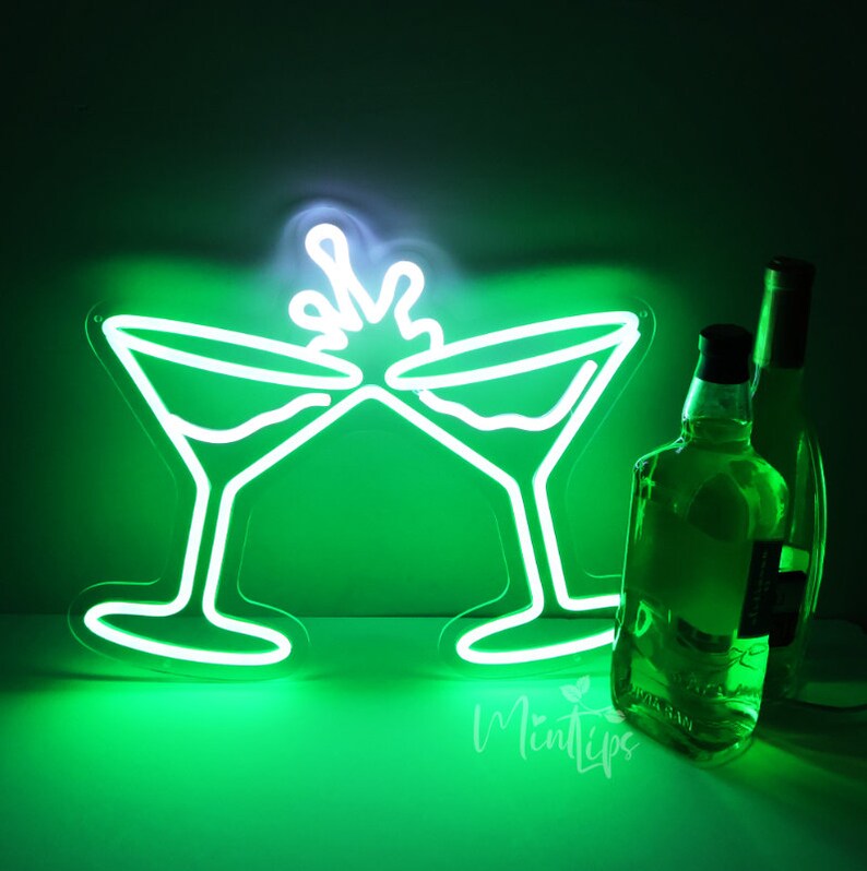 May include: A bright green neon sign depicting two martini glasses clinking together with a splash effect above them. Next to the glowing sign are two clear glass bottles, one positioned slightly behind the other, reflecting the green light.
