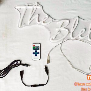 May include: A white neon sign displaying the text The Blelulens, accompanied by a black power adapter, a remote control, a black connector cable, and a 59-inch transparent power line. The components are arranged on a white surface.