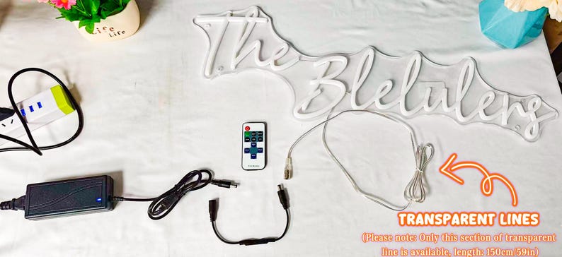 May include: A white neon sign displaying the text The Blelulens, accompanied by a black power adapter, a remote control, a black connector cable, and a 59-inch transparent power line. The components are arranged on a white surface.