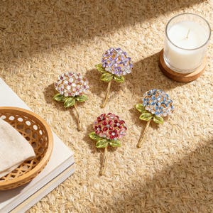 Pode incluir: Quatro ornamentos florais decorativos em tons de rosa, roxo, vermelho e azul. Cada ornamento apresenta pétalas em forma de joia, folhas verdes e uma haste dourada. Uma vela acesa em um pote de vidro está por perto.