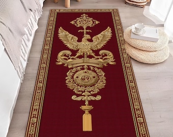May include: A rectangular red rug with a gold ornate design featuring a double-headed eagle, crown, and tassel. The rug has a gold border and is placed on a light-colored wooden floor. The rug's design is symmetrical and detailed.