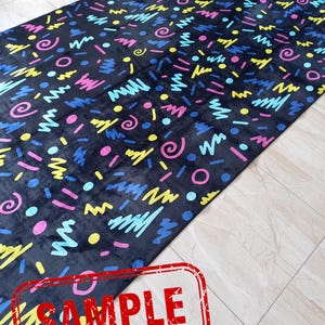 Puede incluir: Una alfombra larga y rectangular de color negro con un patr&oacute;n abstracto y colorido. El dise&ntilde;o presenta formas como garabatos, c&iacute;rculos y puntos en tonos rosa, amarillo, azul y verde. La palabra "SAMPLE" est&aacute; estampada en rojo.