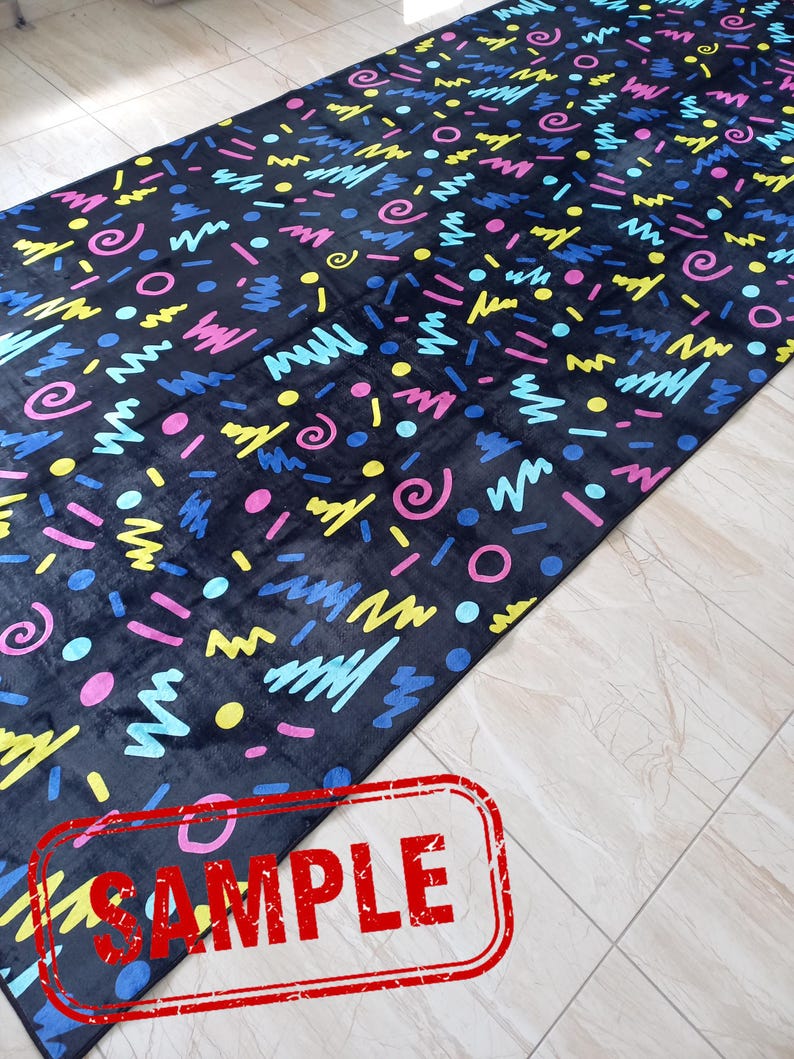 Puede incluir: Una alfombra larga y rectangular de color negro con un patr&oacute;n abstracto y colorido. El dise&ntilde;o presenta formas como garabatos, c&iacute;rculos y puntos en tonos rosa, amarillo, azul y verde. La palabra "SAMPLE" est&aacute; estampada en rojo.