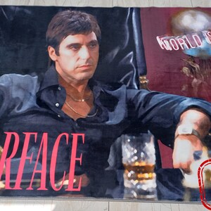 May include: A rug featuring a close-up image of Al Pacino as Tony Montana from the movie Scarface. The text "Scarface" is printed in red on the rug. The background is a red and black color scheme. The text "World is Yours" is printed in white on the rug.