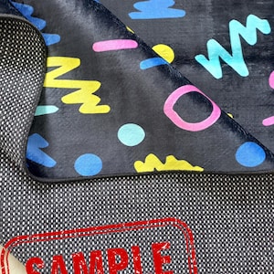 Puede incluir: Una alfombra negra con un colorido dise&ntilde;o geom&eacute;trico estilo a&ntilde;os 80. El dise&ntilde;o incluye formas rosas, azules y amarillas. La alfombra est&aacute; parcialmente doblada, revelando un reverso texturizado. La palabra "SAMPLE" est&aacute; estampada en rojo.