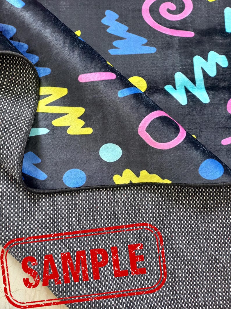 Puede incluir: Una alfombra negra con un colorido dise&ntilde;o geom&eacute;trico estilo a&ntilde;os 80. El dise&ntilde;o incluye formas rosas, azules y amarillas. La alfombra est&aacute; parcialmente doblada, revelando un reverso texturizado. La palabra "SAMPLE" est&aacute; estampada en rojo.