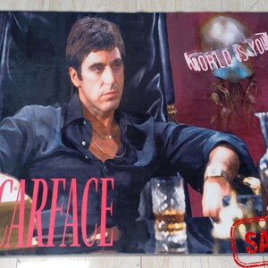 May include: A rug featuring a close-up image of actor Al Pacino as Tony Montana from the movie Scarface. The rug has a red background with the words "World is yours" in white text. The rug also has the word "Scarface" in pink text. The rug has a "Sample" stamp in the bottom right corner.