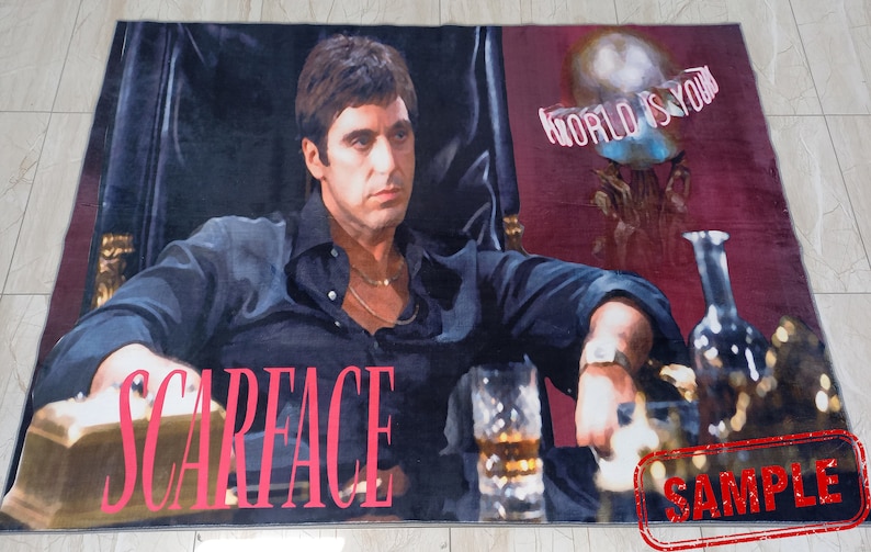 May include: A rug featuring a close-up image of actor Al Pacino as Tony Montana from the movie Scarface. The rug has a red background with the words "World is yours" in white text. The rug also has the word "Scarface" in pink text. The rug has a "Sample" stamp in the bottom right corner.