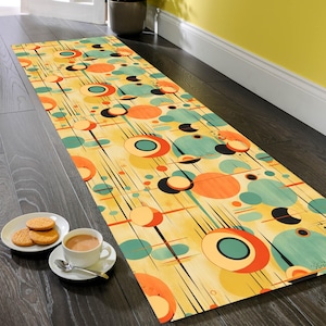 May include: A long, rectangular rug with a mid-century modern design. The rug has a yellow background with orange, teal, and black circular patterns and thin black lines. A cup of tea and biscuits are on the floor next to the rug.