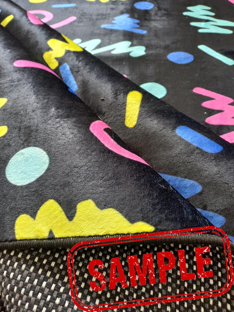 Puede incluir: Alfombra negra con un patr&oacute;n abstracto colorido con formas amarillas, rosas, azules y turquesas. La alfombra tiene un reverso antideslizante. La palabra "SAMPLE" est&aacute; estampada en rojo en la parte inferior.