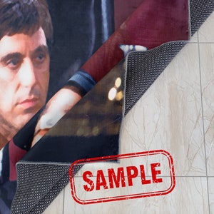 May include: A close-up of a rug with a printed image of a man's face. The rug is folded in half, revealing the back side, which is black with a textured surface. The word "SAMPLE" is printed in red on a white background.