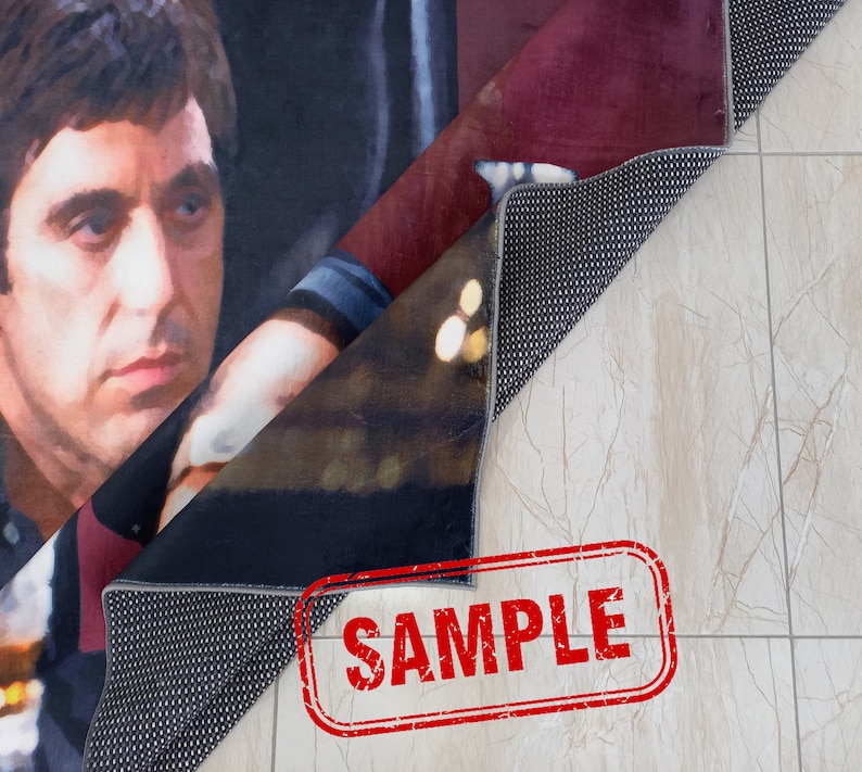 May include: A close-up of a rug with a printed image of a man's face. The rug is folded in half, revealing the back side, which is black with a textured surface. The word "SAMPLE" is printed in red on a white background.