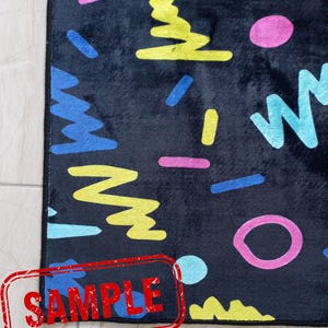 Puede incluir: Una alfombra rectangular negra con un patr&oacute;n geom&eacute;trico colorido. El dise&ntilde;o incluye formas azules, amarillas, rosas y turquesas. La palabra "SAMPLE" est&aacute; estampada en rojo en la esquina inferior izquierda.
