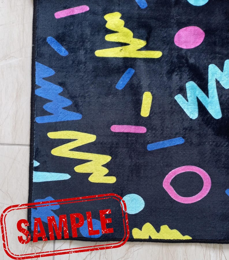 Puede incluir: Una alfombra rectangular negra con un patr&oacute;n geom&eacute;trico colorido. El dise&ntilde;o incluye formas azules, amarillas, rosas y turquesas. La palabra "SAMPLE" est&aacute; estampada en rojo en la esquina inferior izquierda.