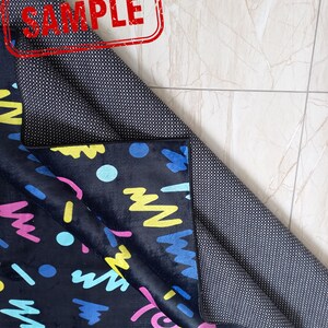 Puede incluir: Una alfombra negra con un dise&ntilde;o abstracto y colorido con formas azules, amarillas, rosas y turquesas. La alfombra tiene un reverso antideslizante. La palabra "SAMPLE" est&aacute; estampada en rojo en la alfombra.