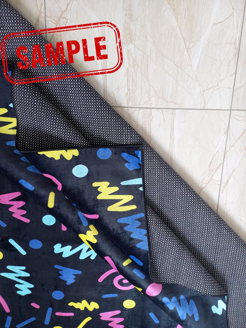 Puede incluir: Una alfombra negra con un dise&ntilde;o abstracto y colorido con formas azules, amarillas, rosas y turquesas. La alfombra tiene un reverso antideslizante. La palabra "SAMPLE" est&aacute; estampada en rojo en la alfombra.