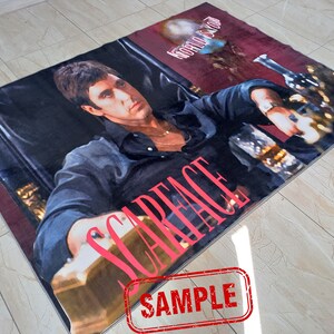 May include: A red and black rug with a picture of a man in a black shirt and a gold chain. The man is sitting in a chair and holding a glass of liquid. The words "Scarface" are printed in red on the rug. The words "World Is Yours" are printed in white on the rug.