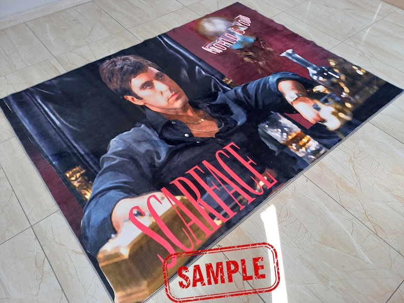 May include: A red and black rug with a picture of a man in a black shirt and a gold chain. The man is sitting in a chair and holding a glass of liquid. The words "Scarface" are printed in red on the rug. The words "World Is Yours" are printed in white on the rug.