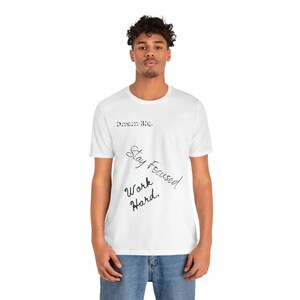 May include: White t-shirt featuring the phrases "Dream Big," "Stay Focused," and "Work Hard" in black script. The shirt has a crew neck design.