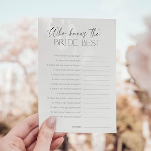 Puede incluir: Una tarjeta blanca con el texto "Who knows the BRIDE BEST" en escritura elegante. La tarjeta presenta una serie de preguntas para completar sobre la novia, perfectas para un juego de despedida de soltera. La tarjeta es sostenida por una persona.