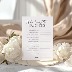 Puede incluir: Una tarjeta blanca con el texto "Who knows the BRIDE BEST" en escritura elegante. La tarjeta presenta preguntas para completar sobre la novia, perfectas para un juego de despedida de soltera. La tarjeta est&aacute; rodeada de flores blancas.