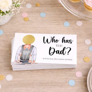 Könnte beinhalten: Ein Stapel Babyparty-Spielkarten mit dem Text "Who has the Dad?" und einer Illustration eines Babys in einer Babytrage. Die Karten sind weiß mit einem goldenen Rubbelkreis. Konfetti und Cupcakes sind im Hintergrund.