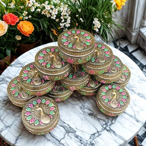 May include: A collection of ornate, round, gold-coloured boxes with peacock designs. Each box features a detailed peacock illustration on the lid, surrounded by pink, green, and gold accents. The boxes are stacked on a marble-patterned surface.