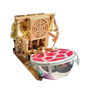 May include: A gold-colored mechanical musical instrument with a drum, bells, and a speaker-like grill. The drum has a red and white geometric pattern. Two drumsticks rest above the drum. The instrument has ornate details and a vintage aesthetic.
