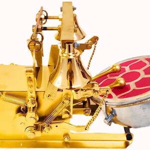May include: A gold-colored musical instrument with a bell, a drum, and a complex system of levers and springs. The drum has a red, patterned surface.