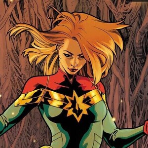 May include: Captain Marvel, a female superhero with blonde hair, wearing a red, blue, and green costume, stands with her arms outstretched, holding glowing orbs of light. She is standing on top of Groot, a tree-like humanoid with a red scarf around his neck. The background is a forest with glowing lights.