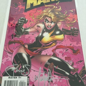 May include: A comic book cover featuring Ms. Marvel in a black and yellow costume with a red sash. The cover is pink with a white background and the title "Ms. Marvel" is in gold letters. The comic book is rated T+ and has a barcode and price information.
