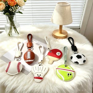 Puede incluir: Colección de llaveros con diseños de deportes y comida. Incluye un béisbol, fútbol americano, gorro de chef, raqueta de ping pong, pelota de tenis y balón de fútbol. Cada llavero tiene una correa de cuero y un cierre de metal.