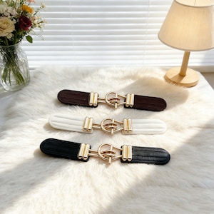 Könnte beinhalten: Drei dekorative Gürtelschnallen in Braun, Weiß und Schwarz. Jede Schnalle hat eine goldfarbene Metallschnalle und liegt auf einer weißen, flauschigen Oberfläche. Die Schnallen sind rechteckig und haben eine gerippte Textur.