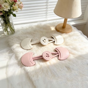 Può includere: Due chiusure a levetta in pelle, una color crema e una rosa, sono esposte su una superficie bianca e soffice. Ogni chiusura presenta un'estremità a forma di mezzaluna e un bottone circolare con cuciture decorative. Ideale per cucito e artigianato.