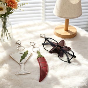 May include: A selection of keyrings and spectacles. The keyrings feature a white martini glass with green olives, and a red chilli pepper with a green stem. Black-framed spectacles rest on a brown star-shaped holder.