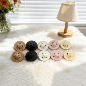 May include: A collection of ten large, round buttons in various colors: tan, black, cream, pink, and pale yellow. Each button has a cross-shaped design in the center. The buttons are arranged on a white, fluffy surface.