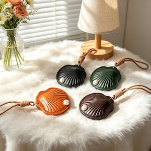 May include: Four shell-shaped leather keychains in black, dark green, tan, and dark brown. Each keychain features a single white pearl accent, a ribbed surface texture, and a tan leather cord loop attached with a wrapped leather knot detail.