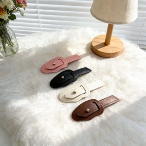 May include: Four decorative belt buckles in pink, black, white, and brown. Each buckle features a rectangular design with a gold-colored button and a matching strap. The buckles are arranged on a white, fluffy surface.