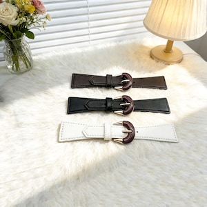 May include: Three leather belts in brown, black, and white, each with a rectangular design and a decorative buckle. The buckles have a gold-colored frame and a dark brown center. The belts are displayed on a white, fluffy surface.