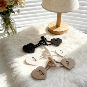 May include: Heart-shaped toggle clasps in black, white, and beige. Each clasp features a heart-shaped button with a letter "E" and a pointed toggle. The clasps are displayed on a fluffy white surface.