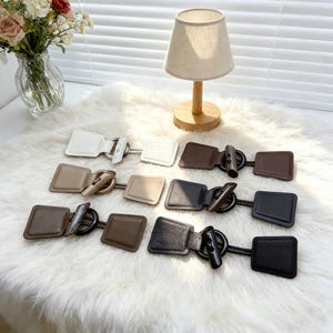 Könnte beinhalten: Eine Auswahl an quadratischen Knebelverschlüssen in Weiß, Beige, Braun und Schwarz. Jeder Verschluss besteht aus einem rechteckigen Lederteil und einem dekorativen Knebel. Die Verschlüsse sind auf einer weißen, flauschigen Oberfläche angeordnet.