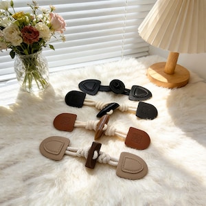 Pode incluir: Uma coleção de fechos de alternância em preto, castanho e bege. Cada fecho apresenta uma peça semelhante a couro com um design decorativo, conectada por uma corda com nós. Os fechos são projetados para casacos, jaquetas ou outras peças de vestuário.