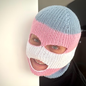 Op de afbeelding: Een gebreide bivakmuts in lichtblauw, roze en wit. Het skimasker bedekt het hoofd en gezicht, met openingen voor de ogen en mond. De kleuren zijn in horizontale strepen gerangschikt.