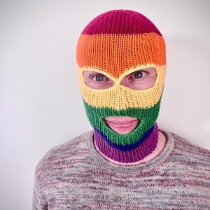Può includere: Un passamontagna lavorato a maglia con una combinazione di colori arcobaleno, con strisce rosse, arancioni, gialle, verdi, blu e viola. La maschera ha aperture per occhi e bocca. La persona indossa un maglione grigio e marrone.