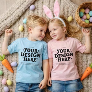 Op de afbeelding: Twee kinderen dragen lichtblauwe en roze t-shirts met de tekst "YOUR DESIGN HERE". Eén kind draagt ​​konijnenoren. Paaseieren en speelgoedwortels liggen om hen heen verspreid. De kinderen liggen op een beige deken.