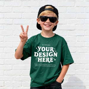 May include: A dark green t-shirt with the text "YOUR DESIGN HERE" in white, worn by a person wearing a black cap and sunglasses. The shirt also has the text "K.C STUDIO" and a line of text that reads "Create your visual/graphic using Photoshop, Canva, or other preferred editing software."