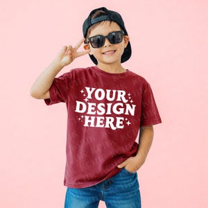 Può includere: Una maglietta bordeaux con la scritta bianca "YOUR DESIGN HERE", indossata da un bambino. Il bambino indossa un berretto nero e occhiali da sole, in posa con il segno della pace. Lo sfondo è rosa.