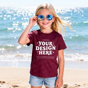 Può includere: Un bambino indossa una maglietta bordeaux con la scritta bianca "YOUR DESIGN HERE", abbinata a pantaloncini di jeans e occhiali da sole rotondi blu. Lo sfondo mostra una spiaggia e l'oceano.