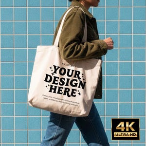 May include: A cream-colored tote bag with the text "YOUR DESIGN HERE" in bold black letters. The bag is carried by a person wearing a green jacket and blue jeans. The background features blue square tiles. The bag also has the text "K.C STUDIO" and "Create your visual/graphic using Photoshop, Canva, or other preferred editing software."